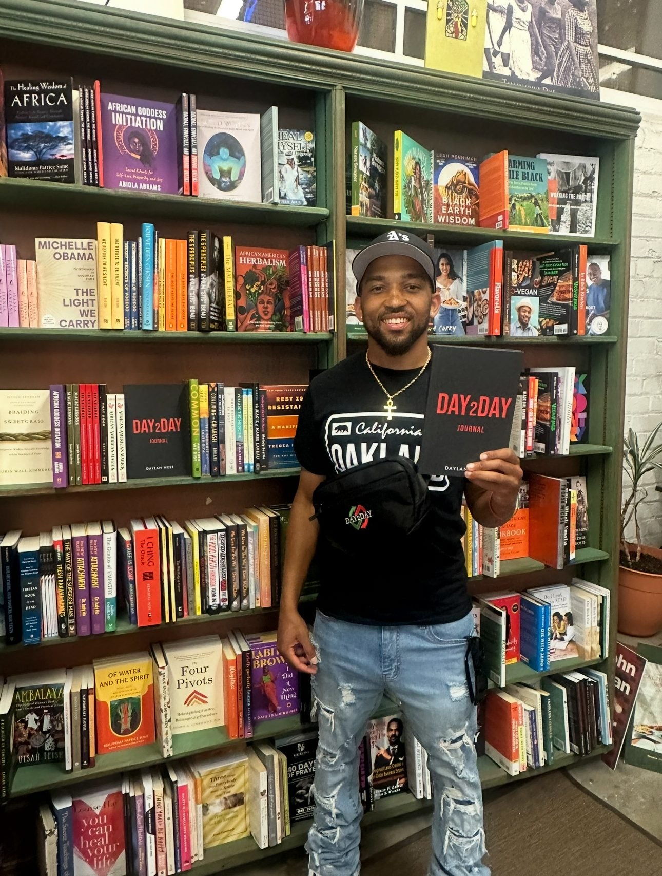 Founder Daylan West holding the Day2Day journal in front of bookshelves at Marcus Books, with the journal also on the shelf